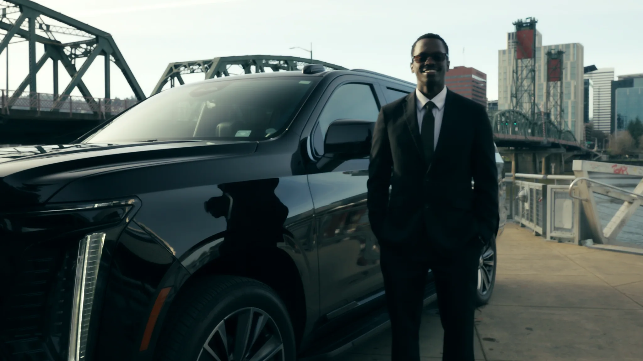 Marquee chauffeur in a suit beside a black Cadillac Escalade with a Portland bridge and skyline behind