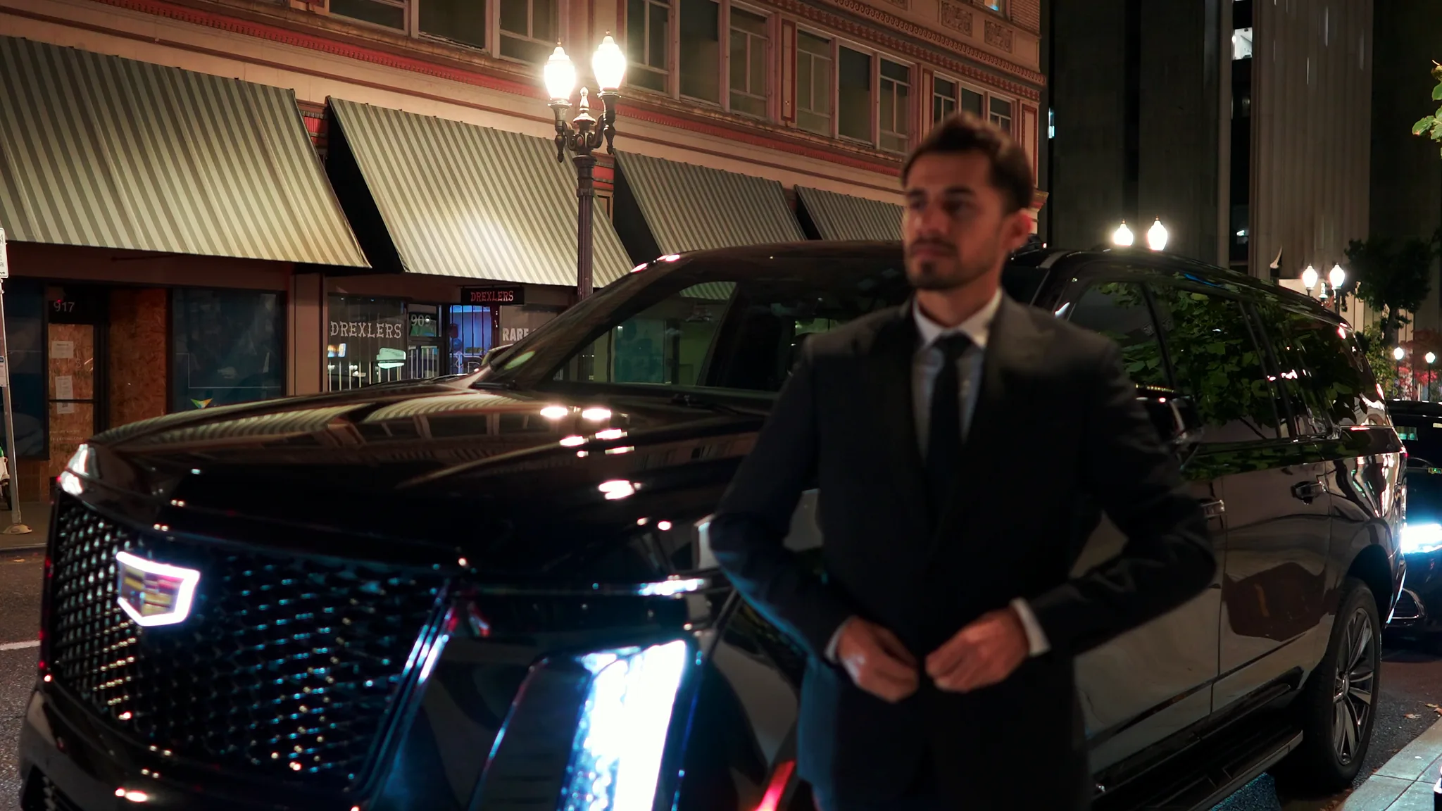 Suited Marquee chauffeur at the rear door of a Cadillac Escalade on a downtown Portland street at night