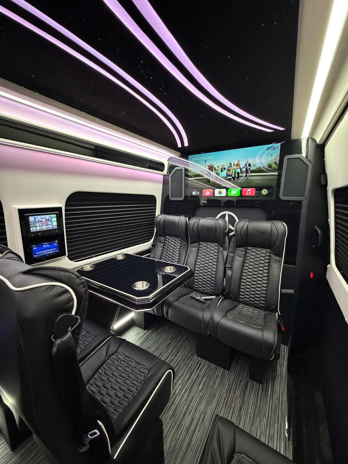 Rear lounge of the Portland Executive Limo: three-seat quilted leather bench under the flat-screen showing the Apple TV home row, gloss-black table with four cup holders, and wall-mounted control screens
