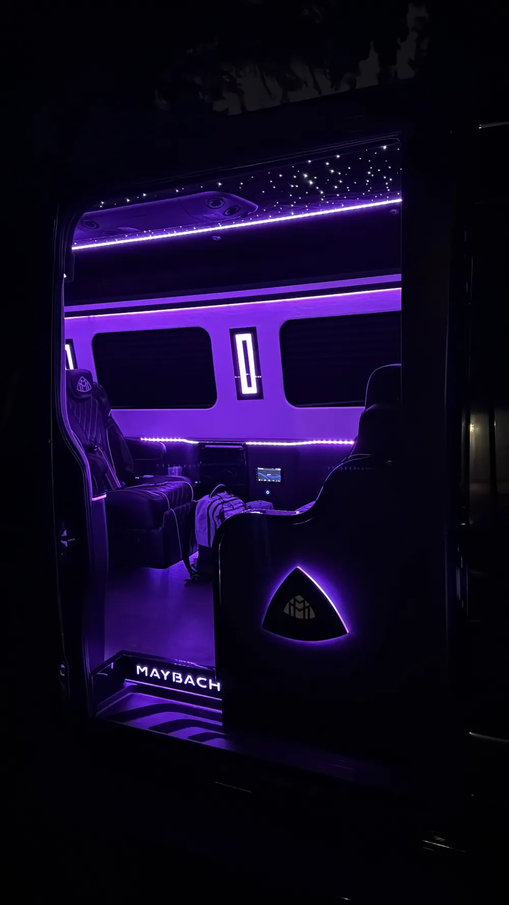 The VIP Lounge Sprinter through its open sliding door at night: illuminated MAYBACH door step, a lit Maybach emblem on the seat side, purple LED, and the starlight ceiling
