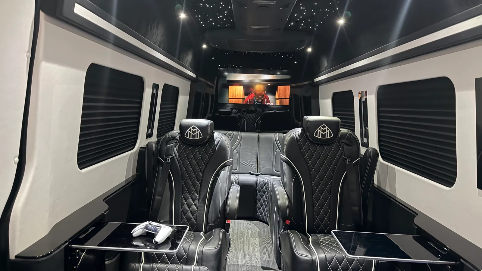VIP Lounge Sprinter cabin seen from the rear bench toward the front: two diamond-quilted captain chairs with Maybach headrest emblems facing rearward, two fold-out gloss-black tables with a game controller on one, window shades, the starlight ceiling, and the front screen