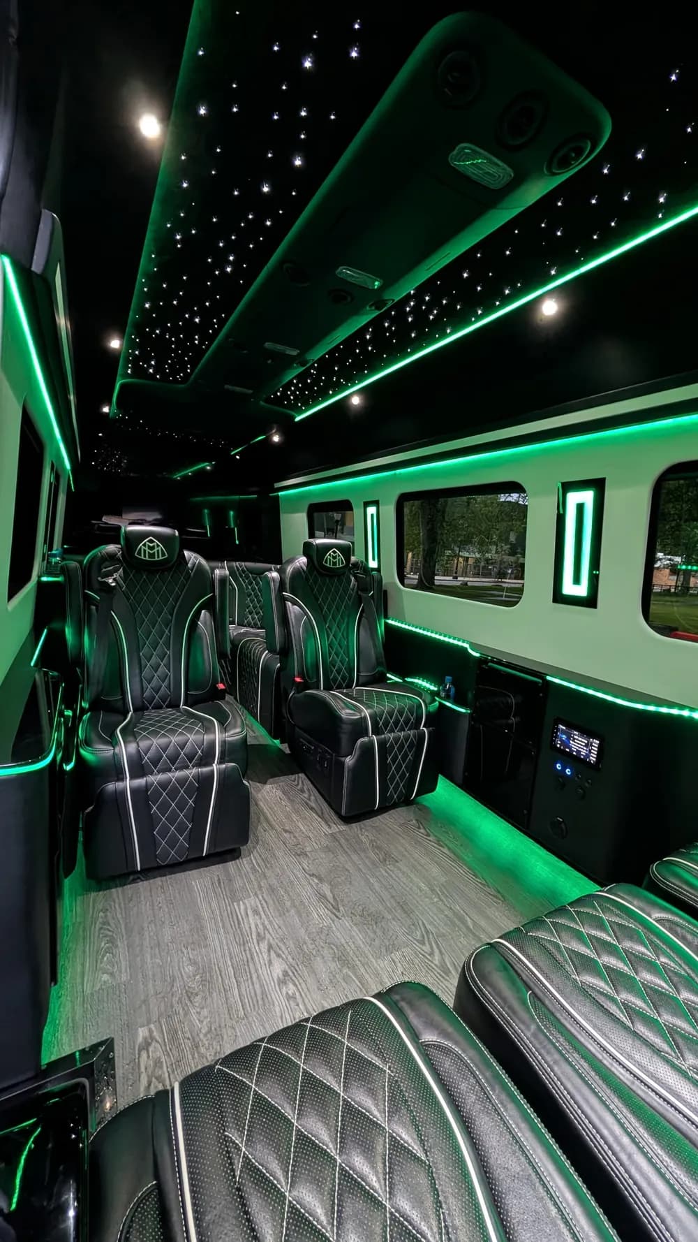VIP Lounge Sprinter cabin under green LED: two captain chairs facing a bench, the black side cabinet with a wall-mounted touchscreen, lit window accents, and floor lighting along the aisle